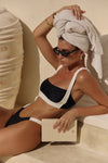 Woman in black bikini sitting on a stone surface with a towel on her head.
